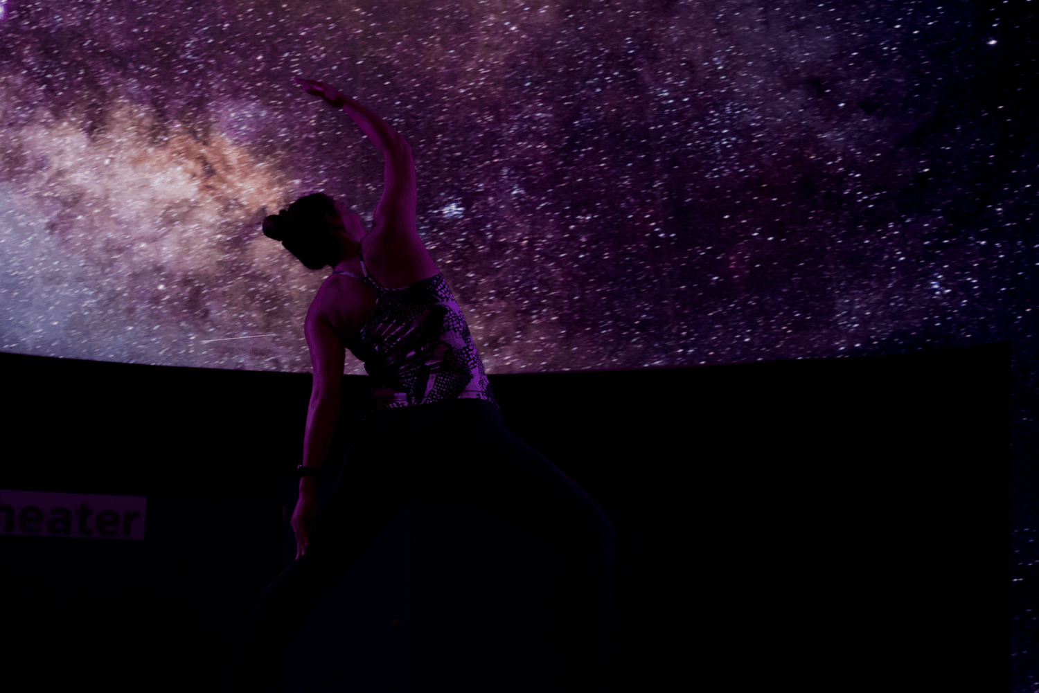 A yoga instructor stretches under the GSK Fulldome Theater. There are stars and nebulas on the screen.