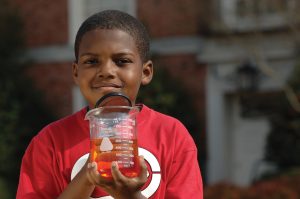 Boy holding beaker filled with orange liquid in one hand and a magnifying glass in the other