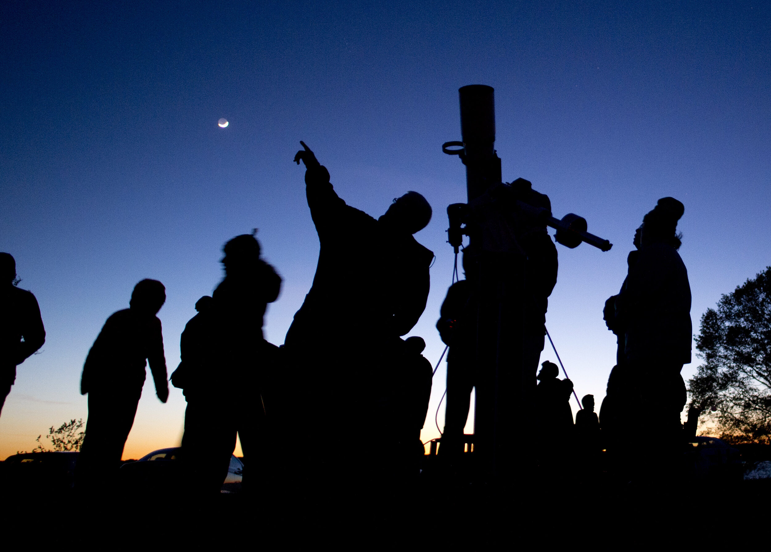 The silhouettes of a group of people standing outside and looking at a telescope. It is dark outside and the sun is setting.
