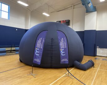 Morehead's Mobile Planetarium deployed in a school gym. Two flags with the Morehead logo and "A gift from Anavi Nagaraj" are deployed outside of the Mobile Planetarium's entrance.