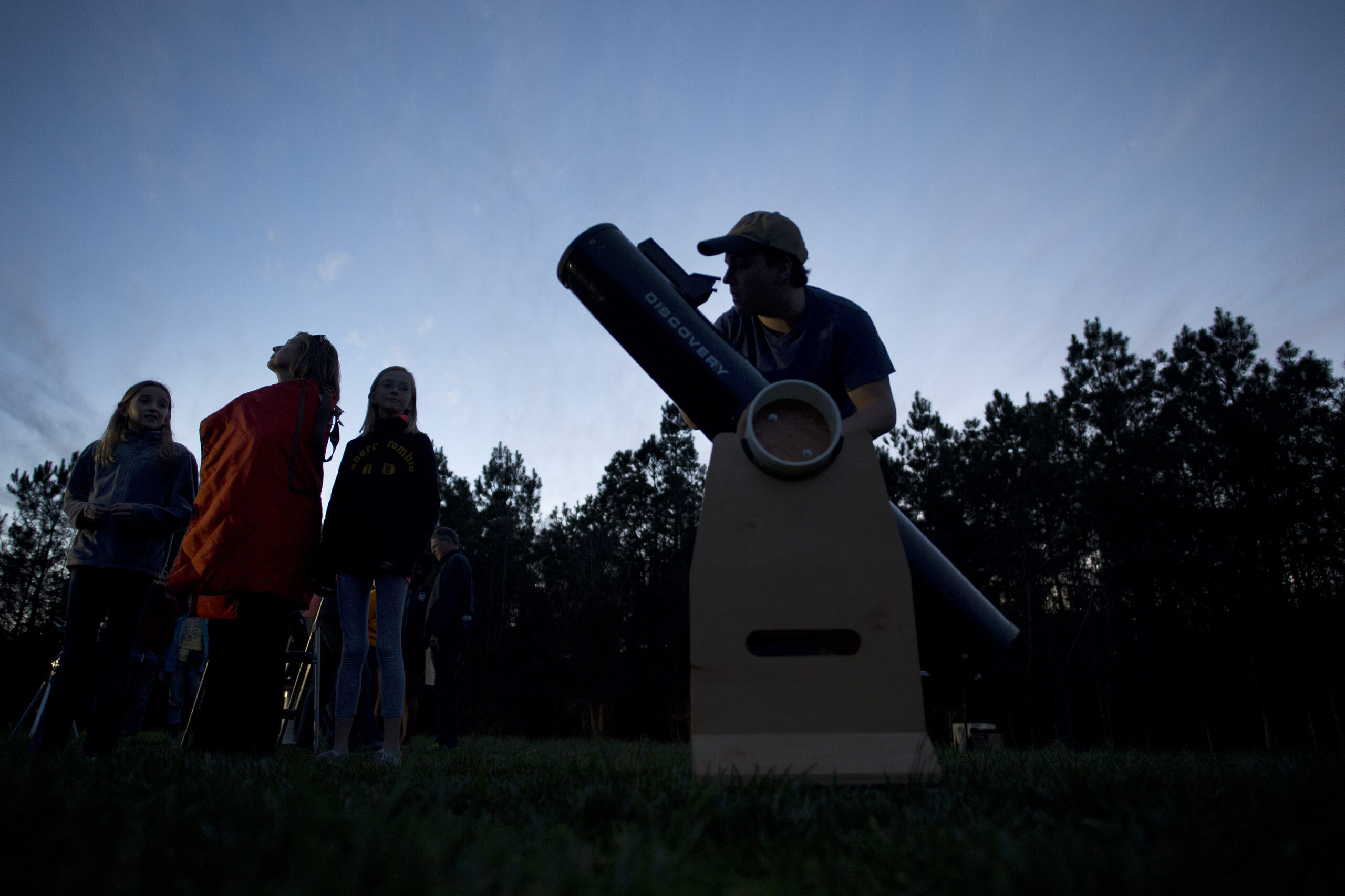 A group of people look at the sky. One person looks through a telescope. It is bright outside, but the sun is setting.