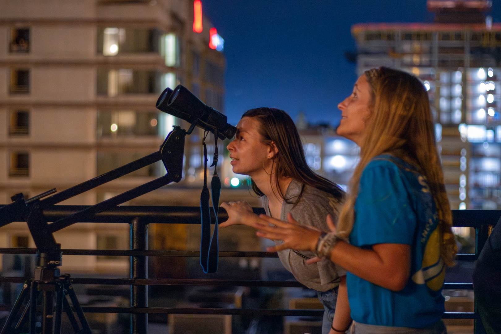 A Morehead educator speaks with a guest looking through binoculars. In the background is the Durham skyline.