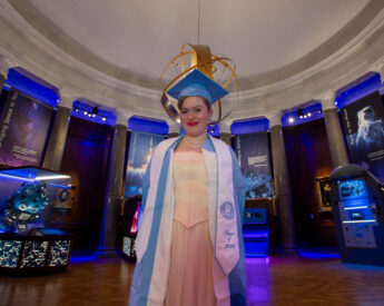 Noelle Keister stands in a grad cap and gown in the Morehead Planetarium and Science Center Gateway Gallery.