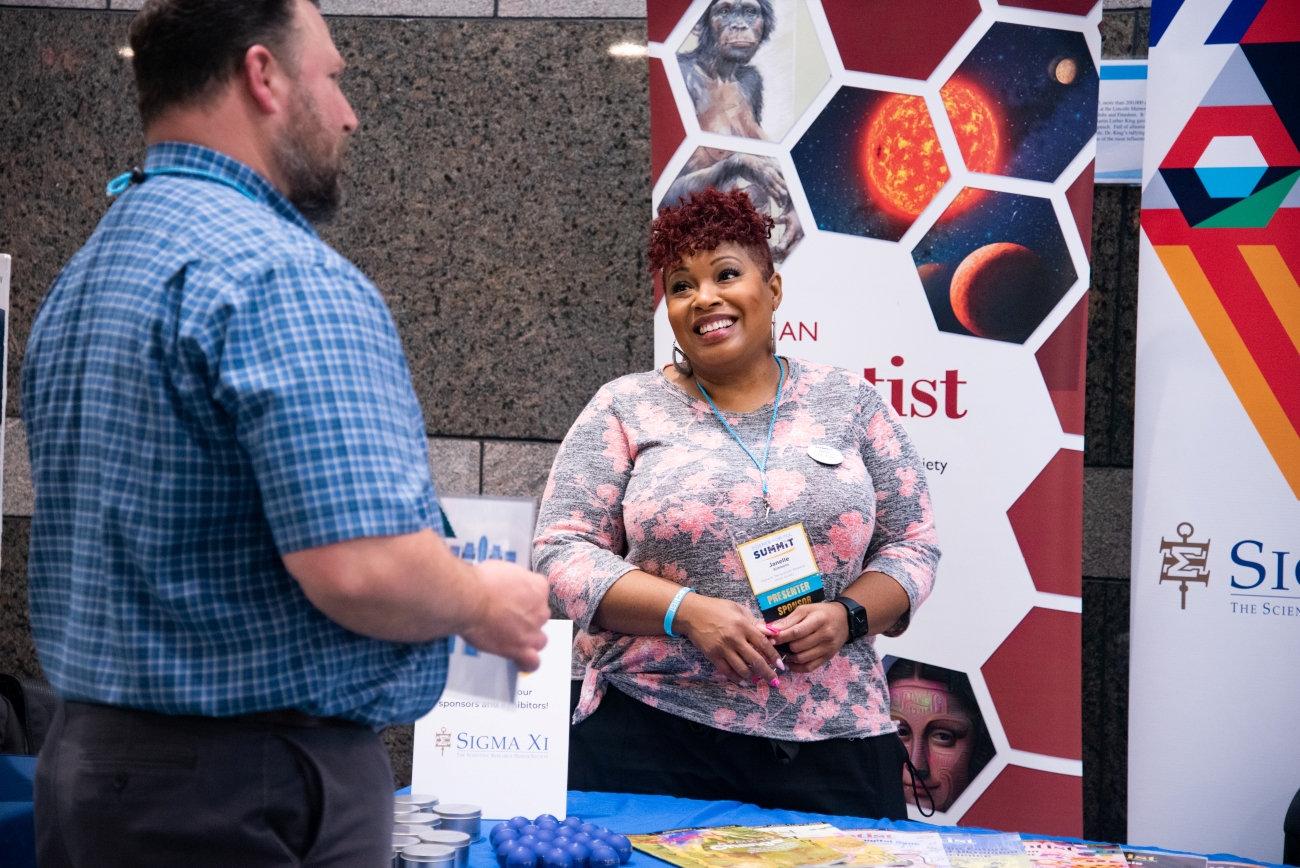 Two professionals talk at a booth. In the background is a science banner.