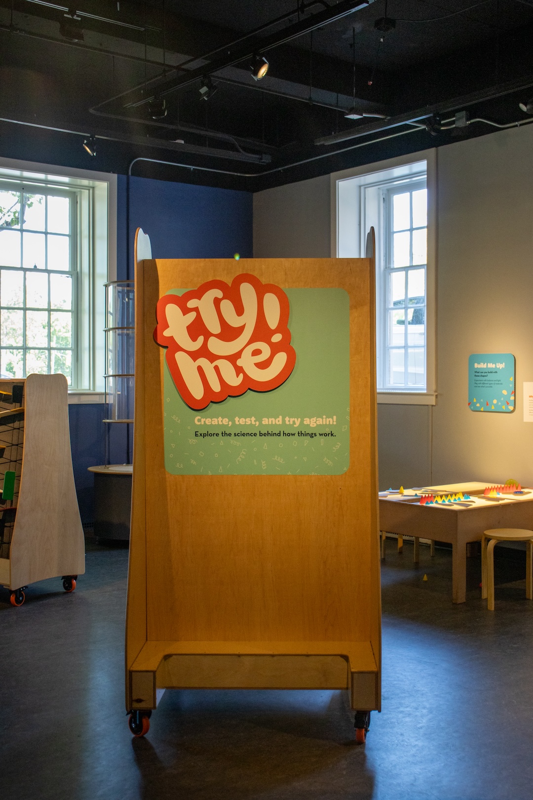 The Try Me! exhibit sign on a bright day. In the background, there are tables and other science materials.