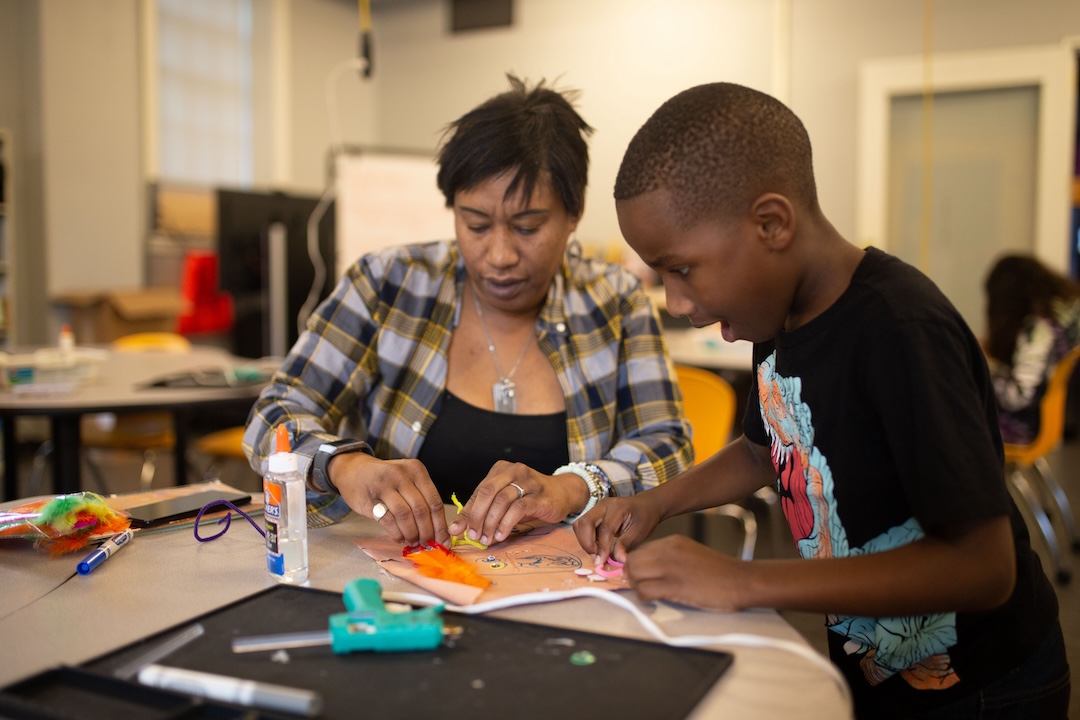 A child and adult sit at a table in the Launch Lab. On the table, there is glue, paper, and a hot glue gun.