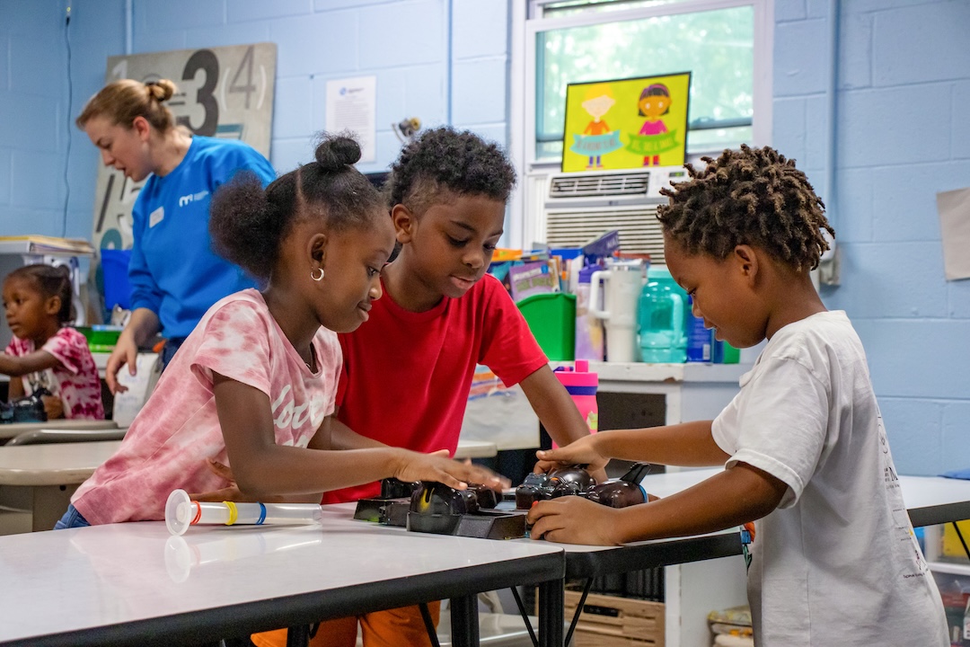 Students stand around a desk in a classroom. They play a rendition of Hungry Hungry Hippos focused on modeling star formation.