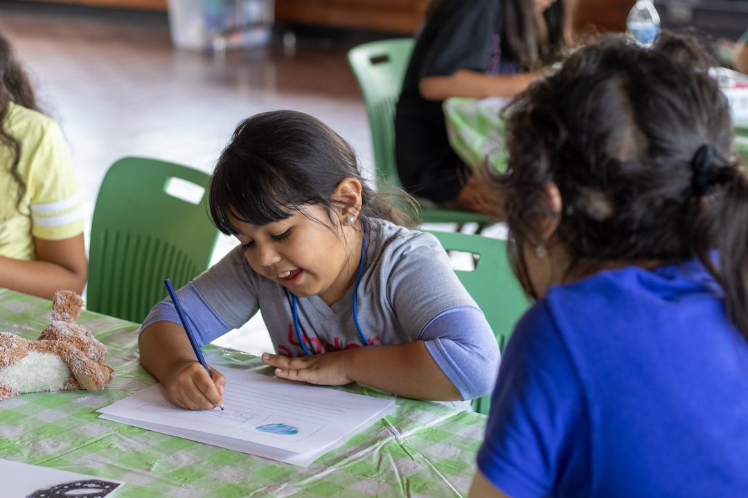 A child sits at a table while using a colored pencil on a sheet of paper.