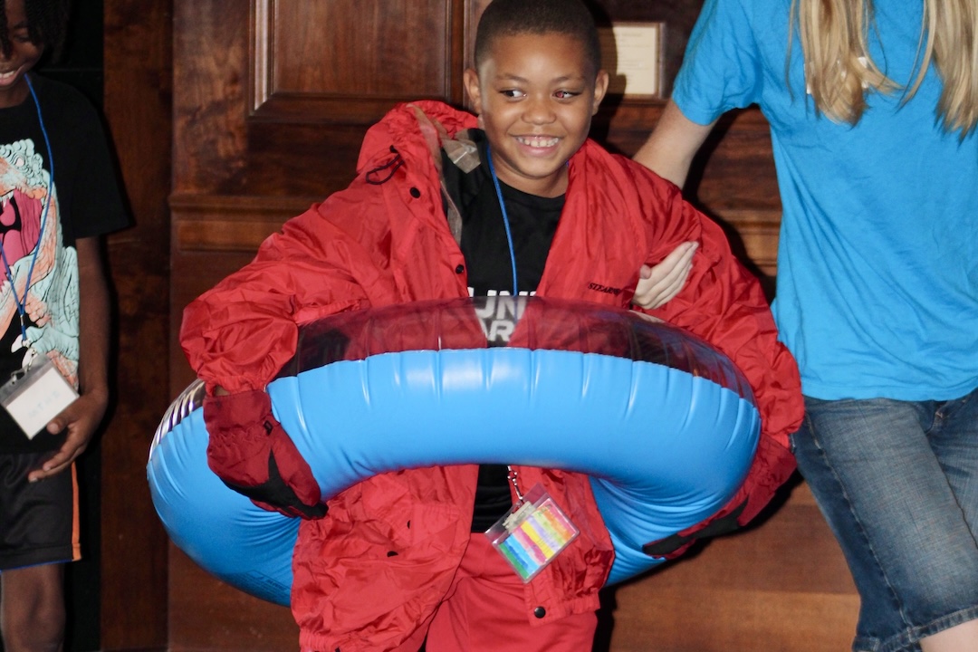 A child wears an inflatable tube and a red coverall suit with gloves. In motion, the child smiles.