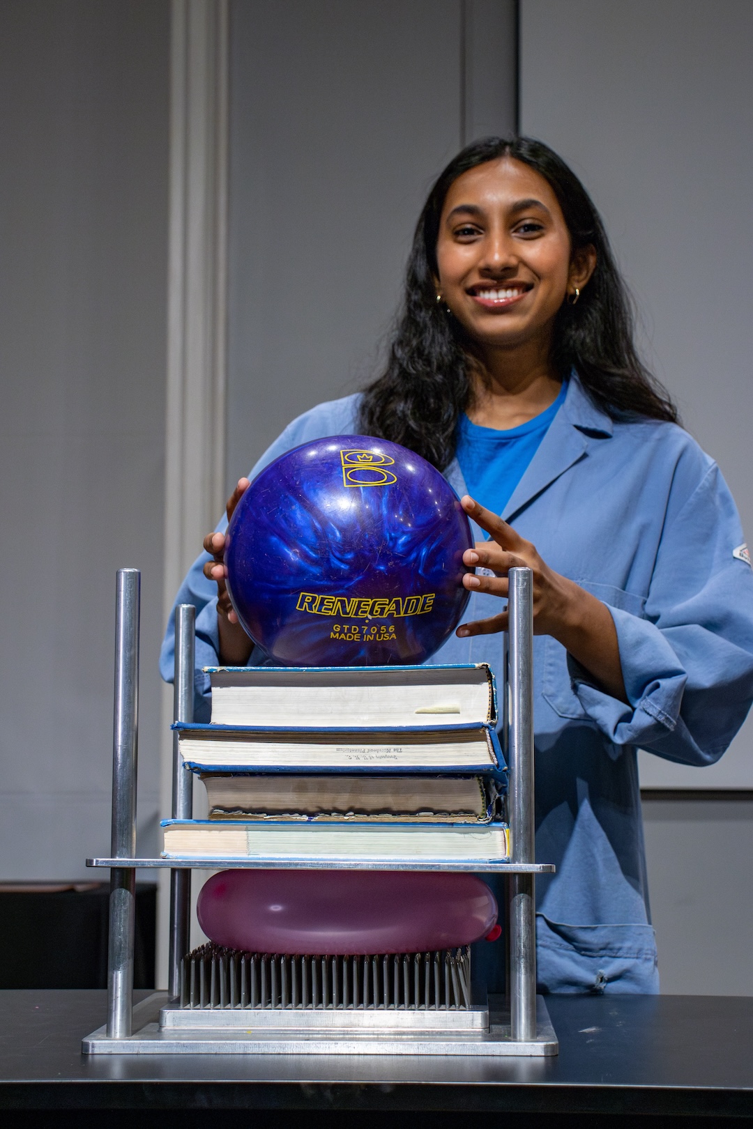 On a bed of nails is a balloon and four large textbooks. A Morehead student employee holds a bowling ball over the other objects to model weight distribution. 