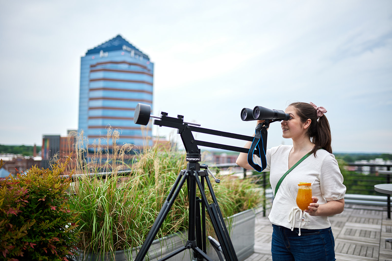 A woman stands on a rooftop while holding a cocktail. She looks through a pair of astronomy binoculars.