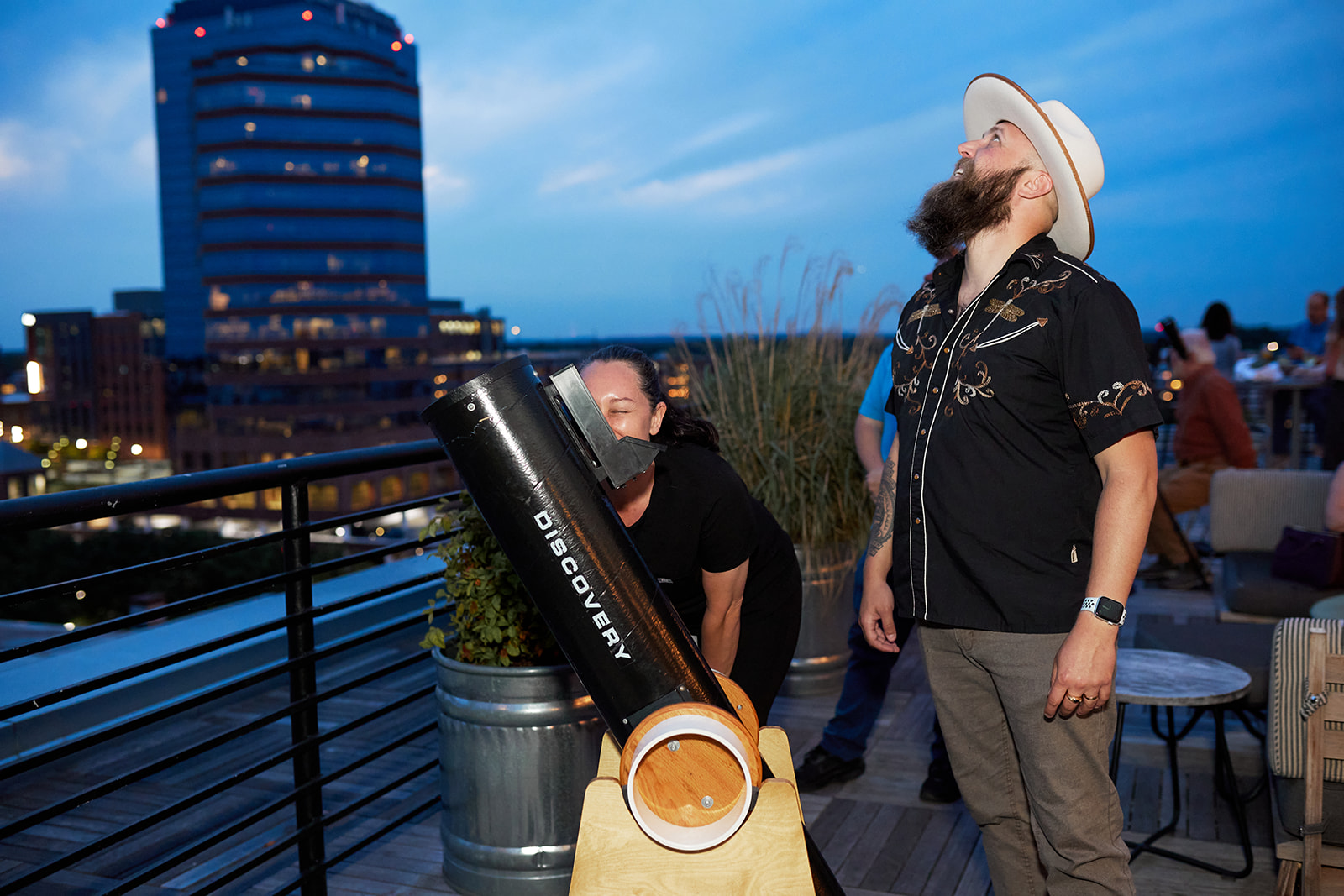 Two people stand on a decorated rooftop. One looks through a telescope while the other looks up at the sky.