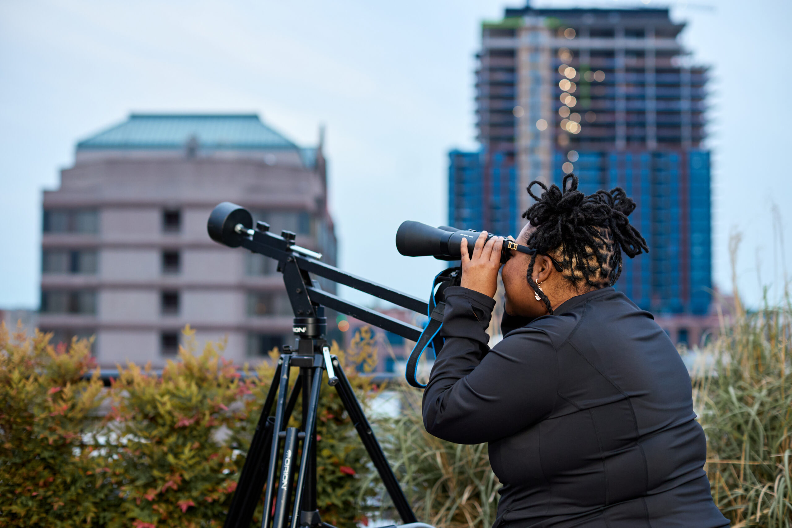 A person with braided space buns looks through a pair of binoculars. In the background is part of the Durham city skyline.