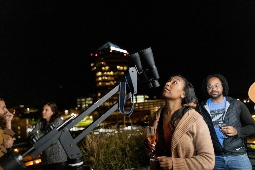 A woman looks up at the sky while holding a champagne glass. In front of her is a pair of binoculars.