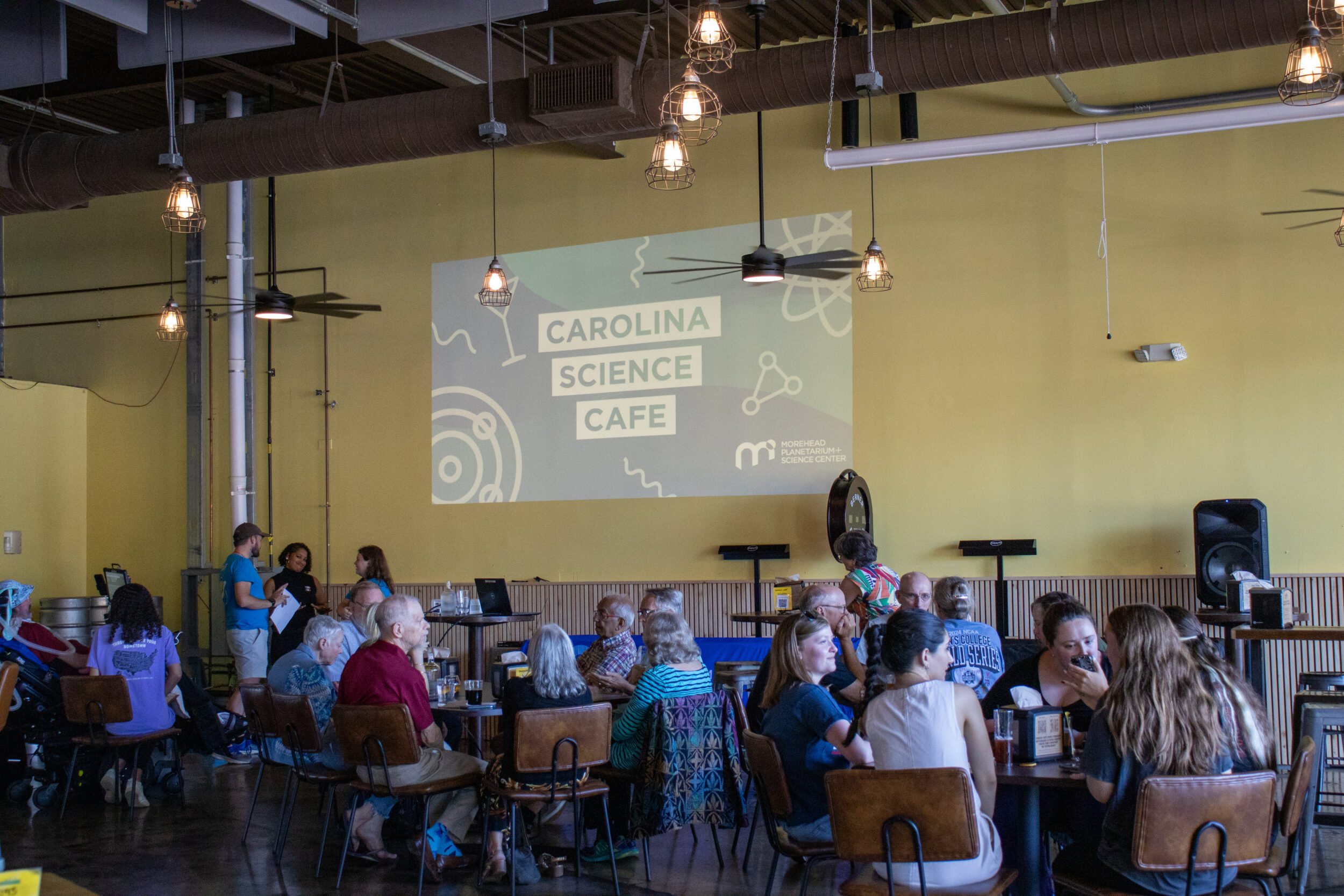 A crowd seated at multiple tables. "Carolina Science Cafe" is printed on the wall.