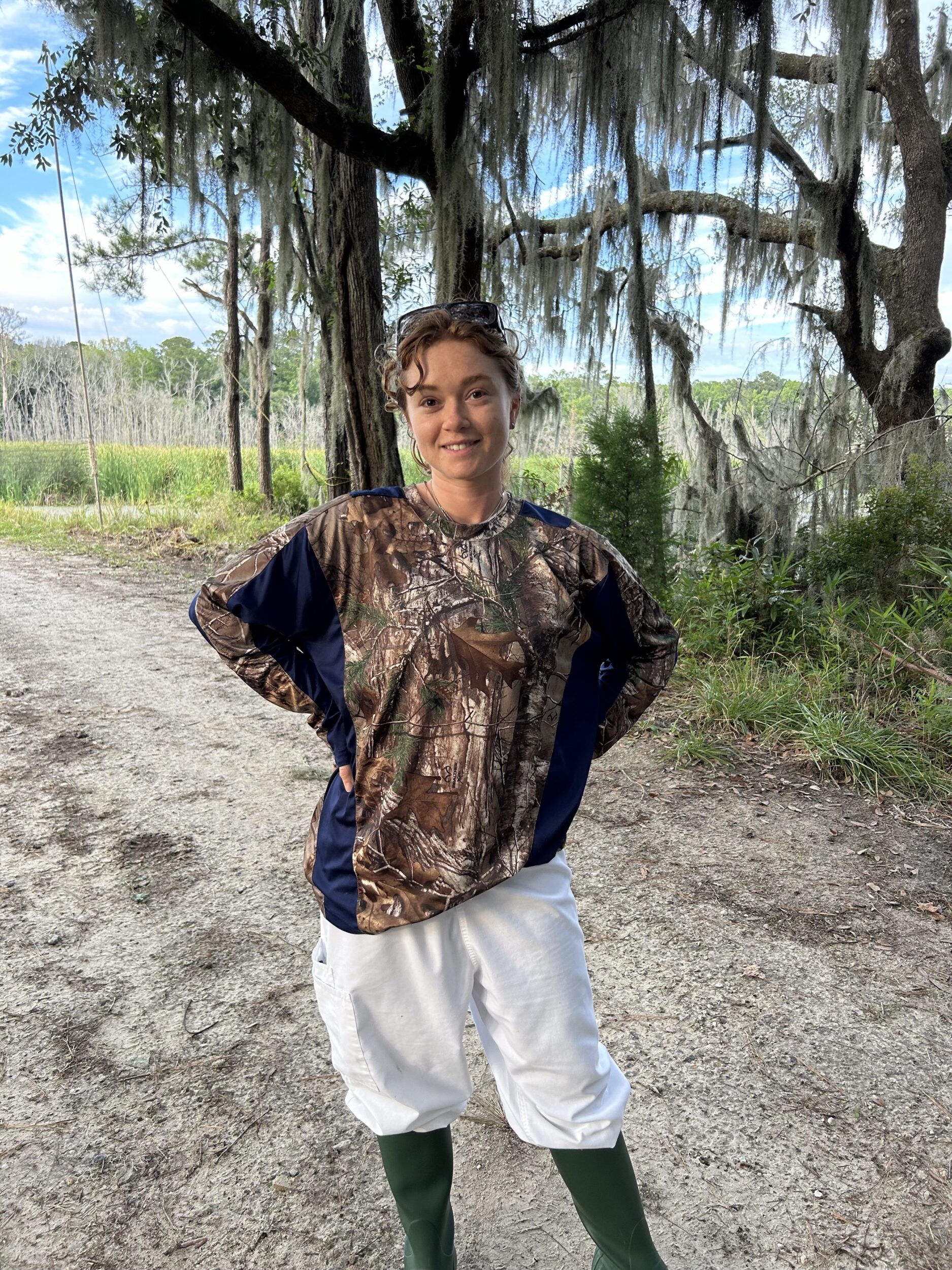 Madeleine Popkin poses in wetlands while wearing a camo shirt and tall black boots.