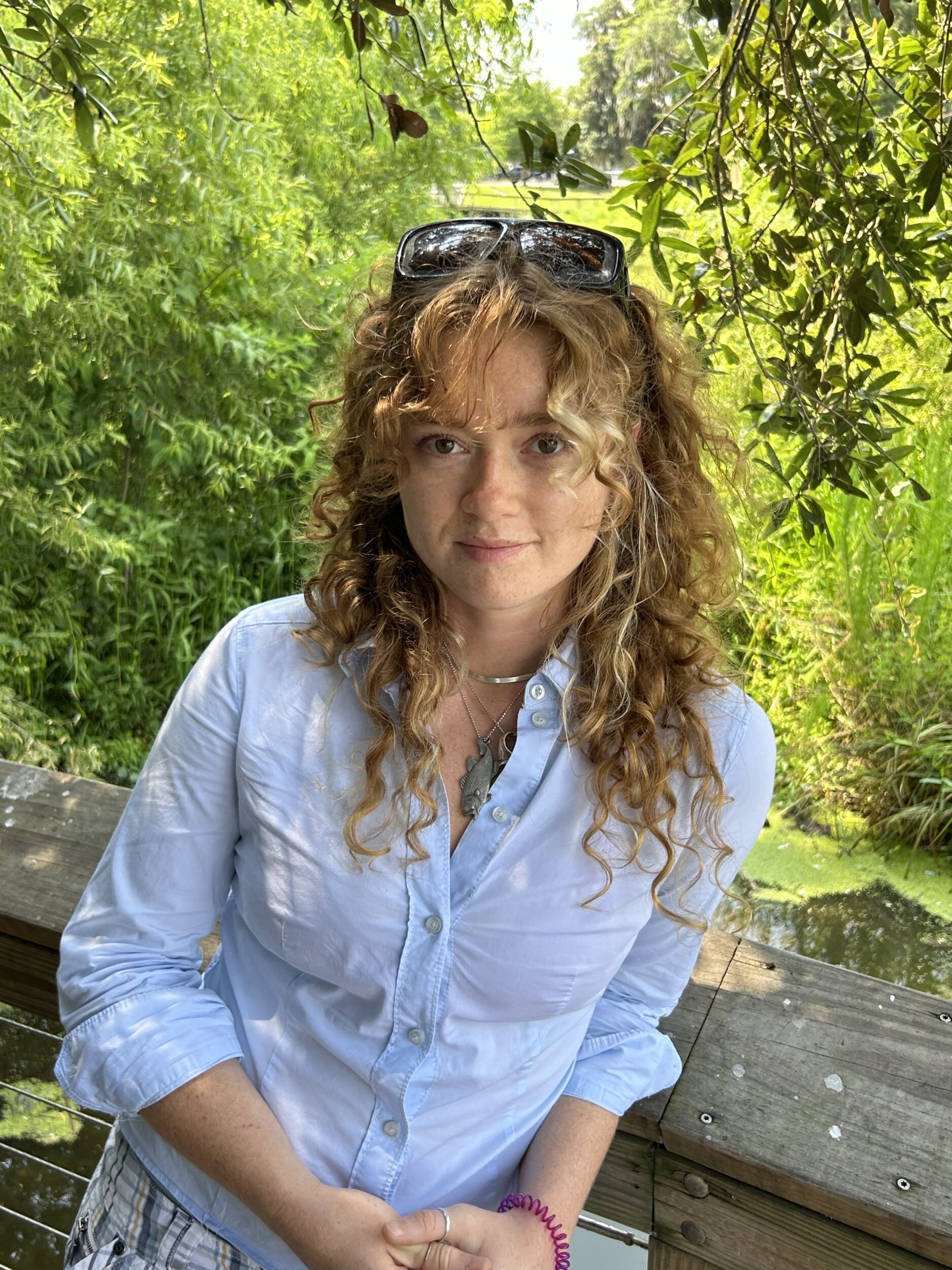 Madeleine Popkin smiles in a blue button-down shirt. She has sunglasses on her head and stands in front of greenery.