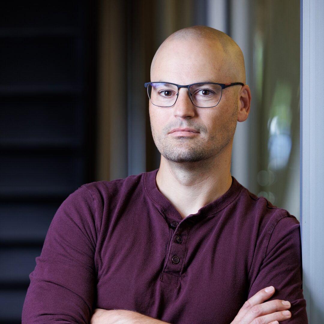 Dr. Jonathan Campbell leans against a wall while cross his arms. He wears glasses and a burgundy shirt.