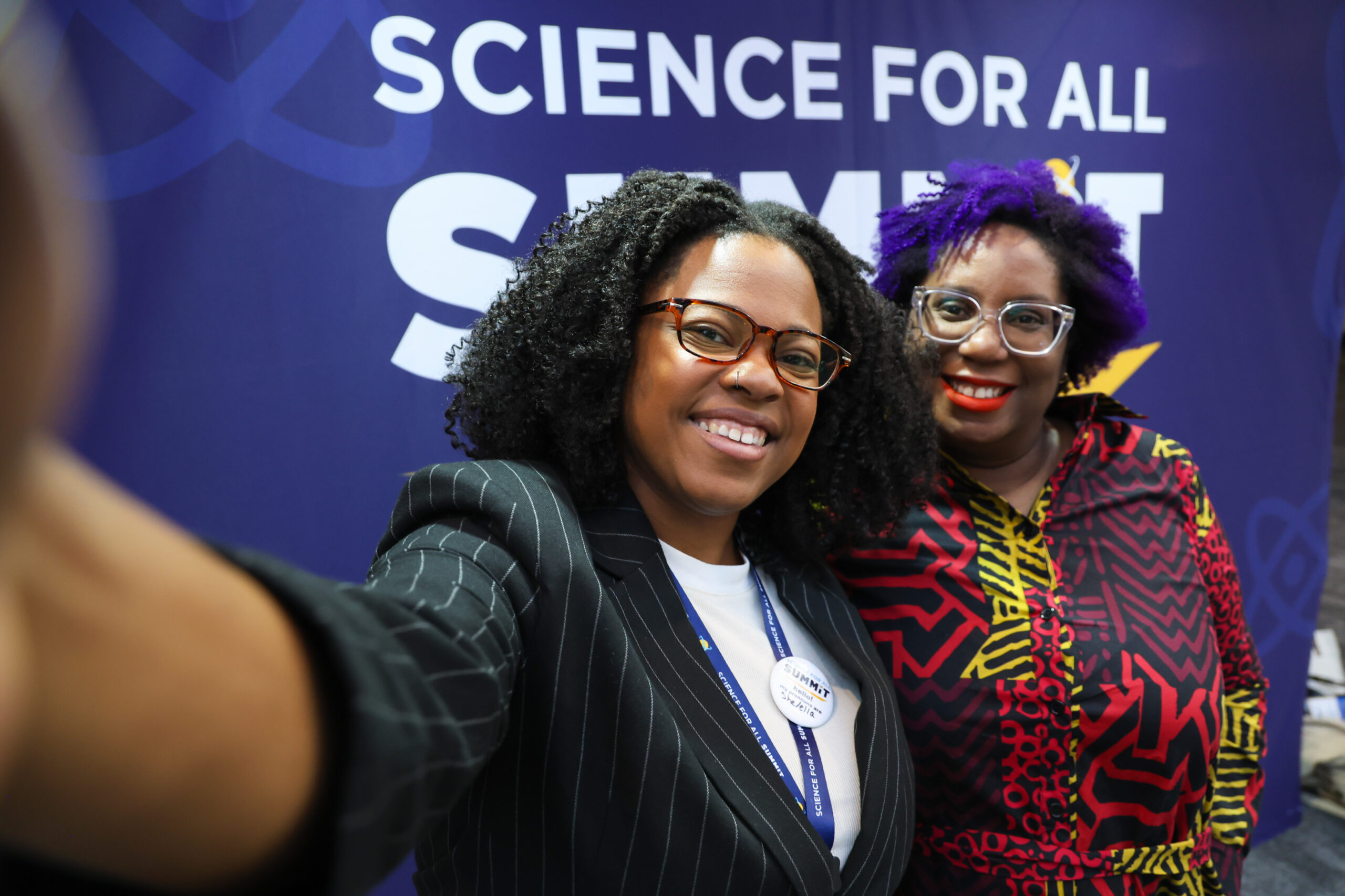 Two attendees with curly hair smile and take a selfie in front of a Science for All Summit backdrop.