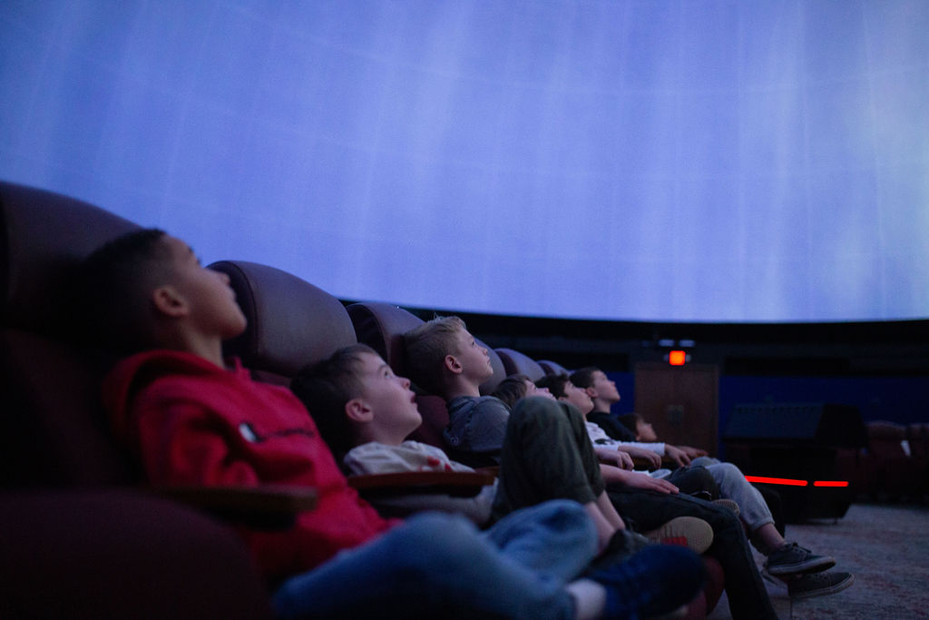 A group of children sit next to each other in the fulldome theater. Shades of blue are projected on the dome. 