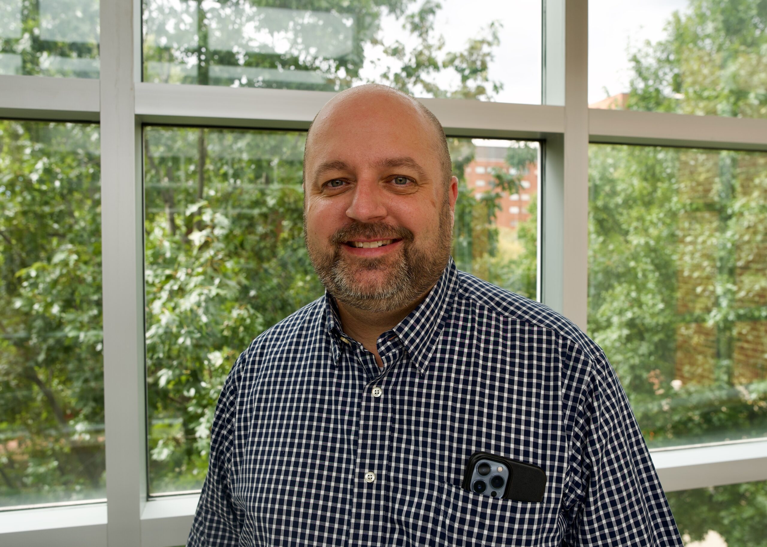 Dr. Jared H. Bowden smiles in front of a window overlooking trees. He wears a checkered button-down shirt.
