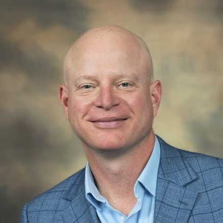 Dr. Jason Roberts smiles against a neutral background. He wears a blue suit with a light blue shirt and handkerchief.